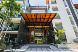 A modern building entrance with a covered walkway, glass doors, and surrounding greenery. - At The Tree Sea View 1BR Walk To Rawai Beach (Rawai)