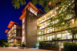 Et hotel med flere etager, et tydeligt skilt med 'AT THE TREE', store vinduer og planter ude.