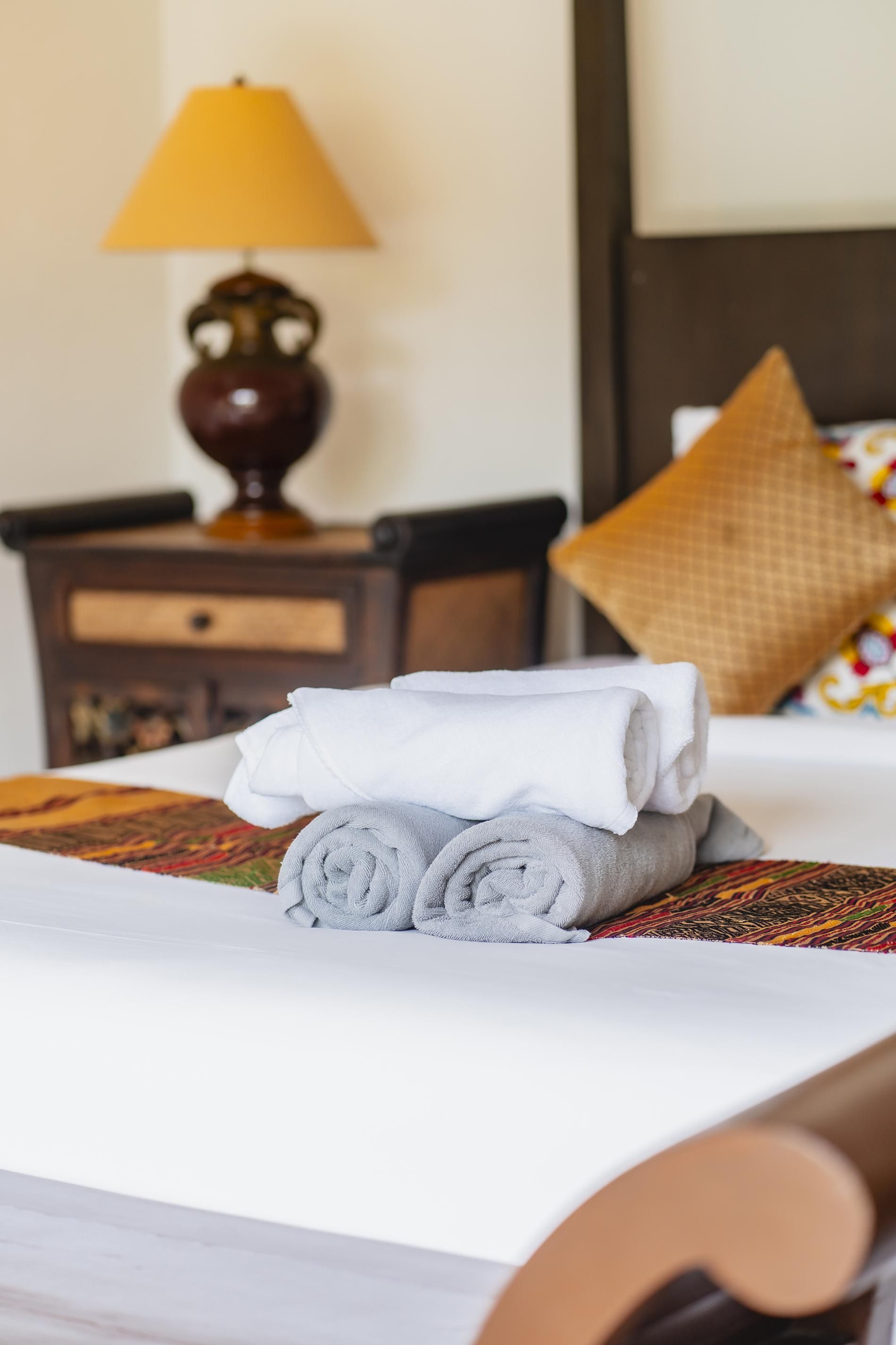 A neatly made bed with rolled towels, a patterned bedspread, a wooden nightstand with a lamp, and a pillow with a geometric design.