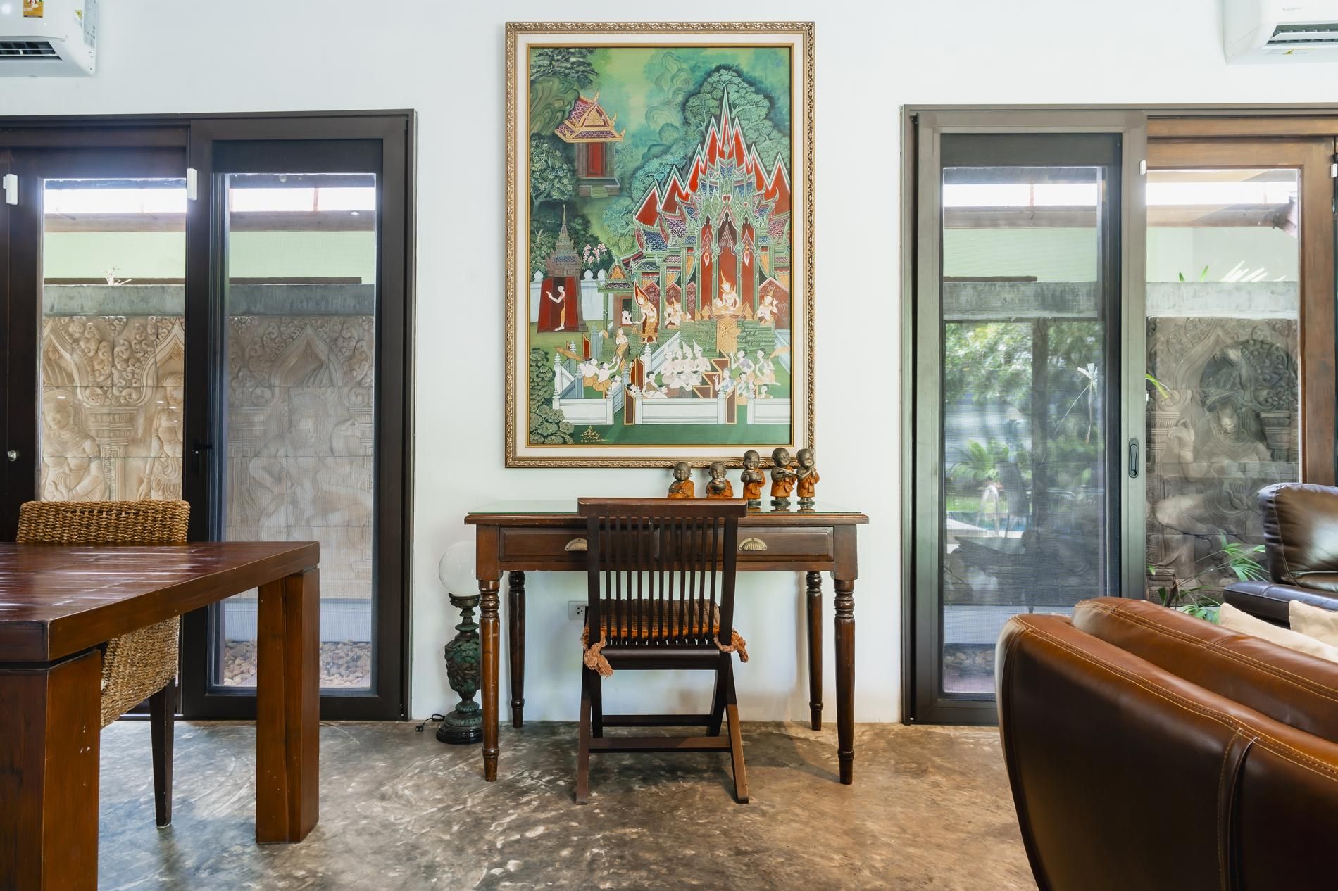 Ein Zimmer mit traditionellem thailändischen Wandgemälde, einem Holztisch, einem Ledersofa und einem gewebten Korb.