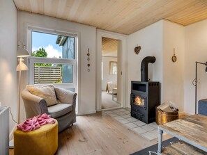 A cozy room with a sofa, a small ottoman, a table, a wood-burning stove, and a window with blinds. - "Helemine" - 800m from the sea by Interhome (Blokhus)