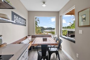 A modern kitchen with a wooden table and chairs, a built-in bench, and a view of a lake through large windows. - Modern 2-bedroom Container Home with beautiful river views (Canby)