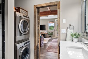 A laundry area with stacked washers and dryers, a wooden door leading to a living space with a TV, a plant, and a view of greenery. - Modern 2-bedroom Container Home with beautiful river views (Canby)