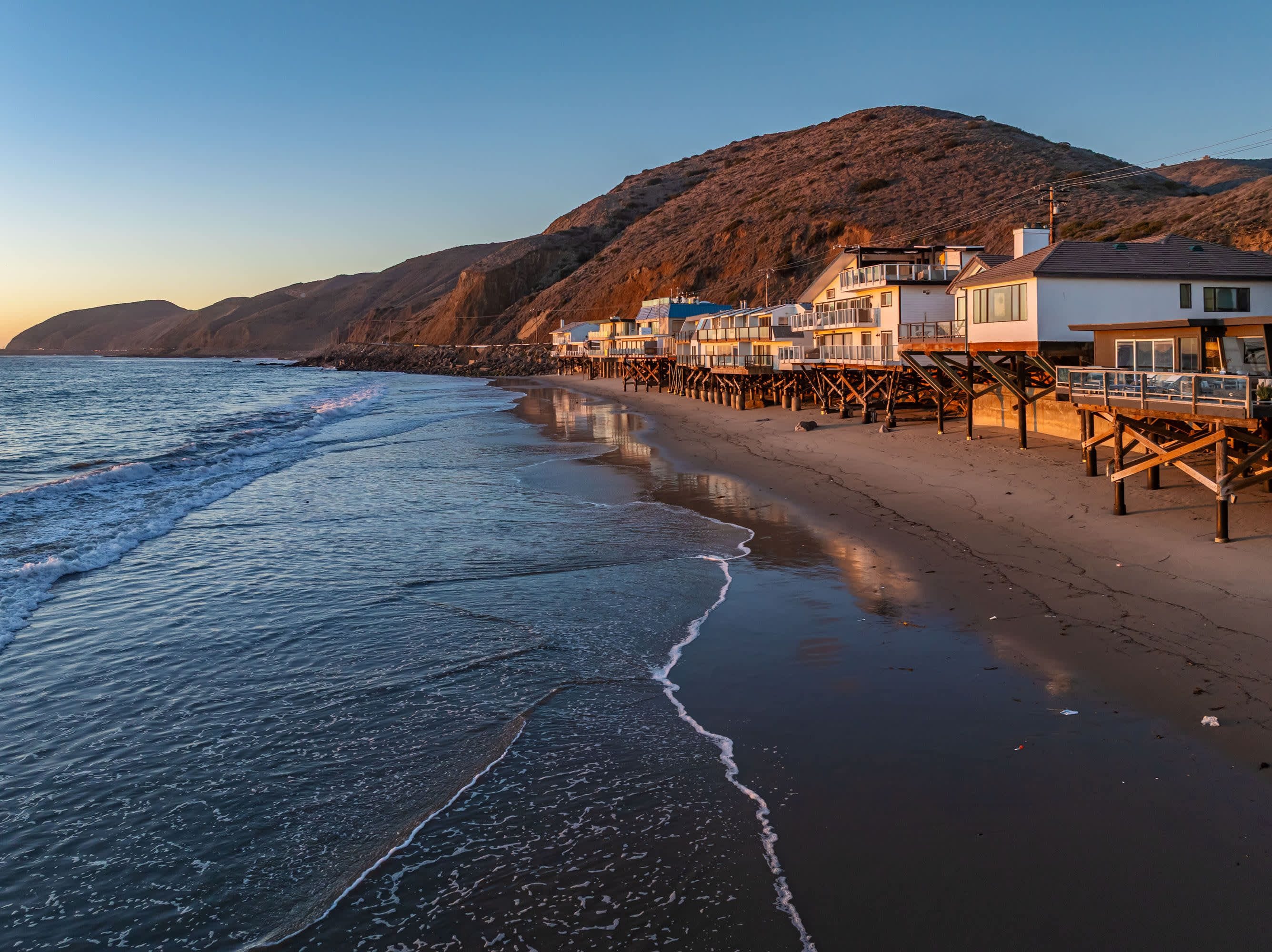 A coastal hotel with multiple buildings on stilts, a beach, and a mountainous backdrop.
