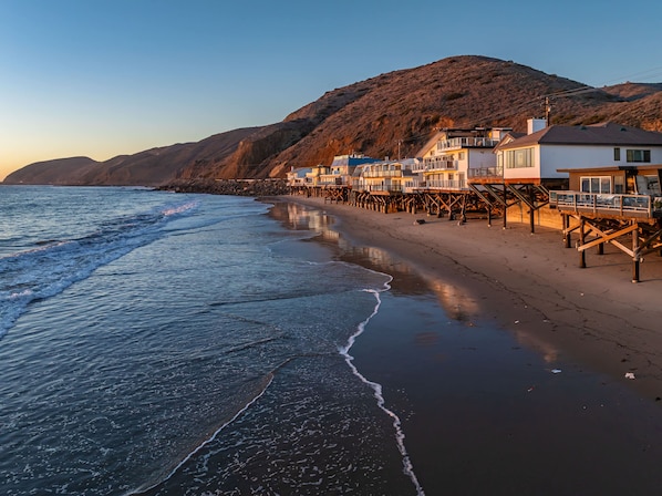 A coastal hotel with multiple buildings on stilts, a beach, and a mountainous backdrop.