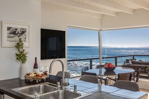 A modern kitchen with a large window overlooking the sea, a dining area with a table and chairs, and a wine bottle on the counter.