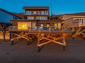 A modern two-story house with a balcony, built on stilts over a sandy area.