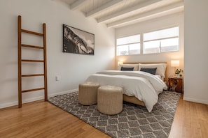 A bedroom with a bed, two ottomans, a ladder, a painting, and a window.