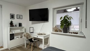 A room with a white desk, a television mounted on the wall, a window with a plant, and a small clock on the windowsill. - Guest Rooms Eifelgluck in Pelm (Pelm)