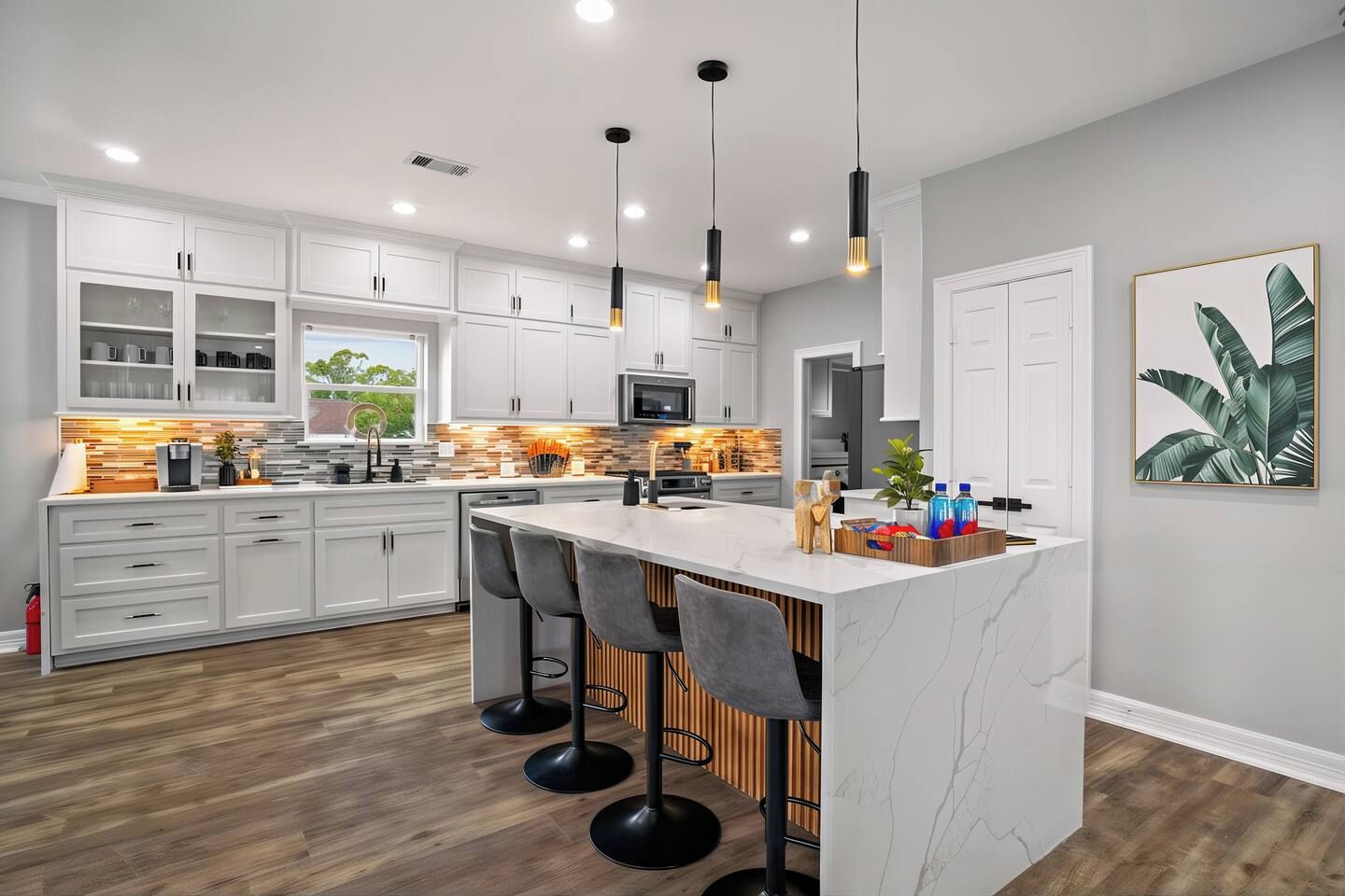A modern kitchen with white cabinets, a marble island, bar stools, pendant lighting, and a large window.