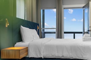 Una habitación de hotel moderna con una cama grande, una mesita de noche de madera y vistas al mar a través de amplias ventanas.