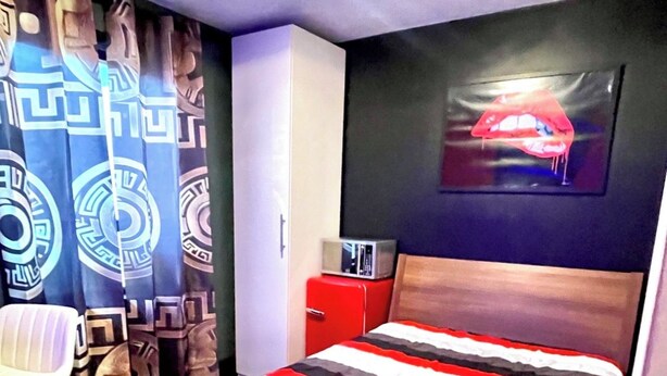 Un dormitorio con una cama, una silla, un armario rojo y una pared con un cuadro.