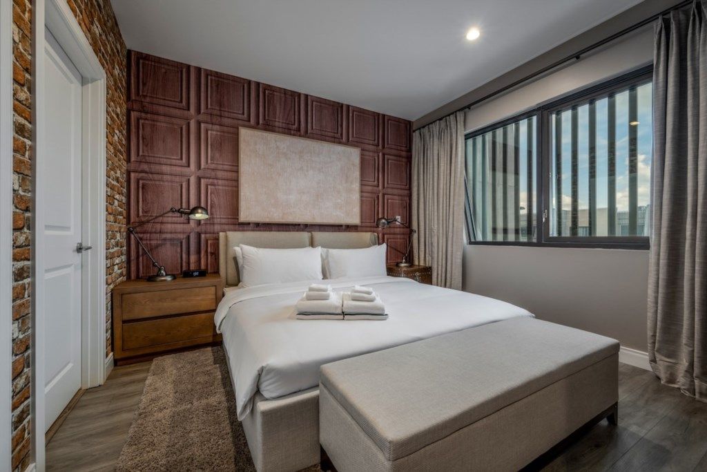 A modern hotel room with a large bed, a wooden accent wall, a nightstand with a lamp, and a view of the city through large windows.