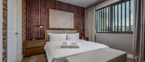 A modern hotel room with a large bed, a wooden accent wall, a nightstand with a lamp, and a view of the city through large windows.