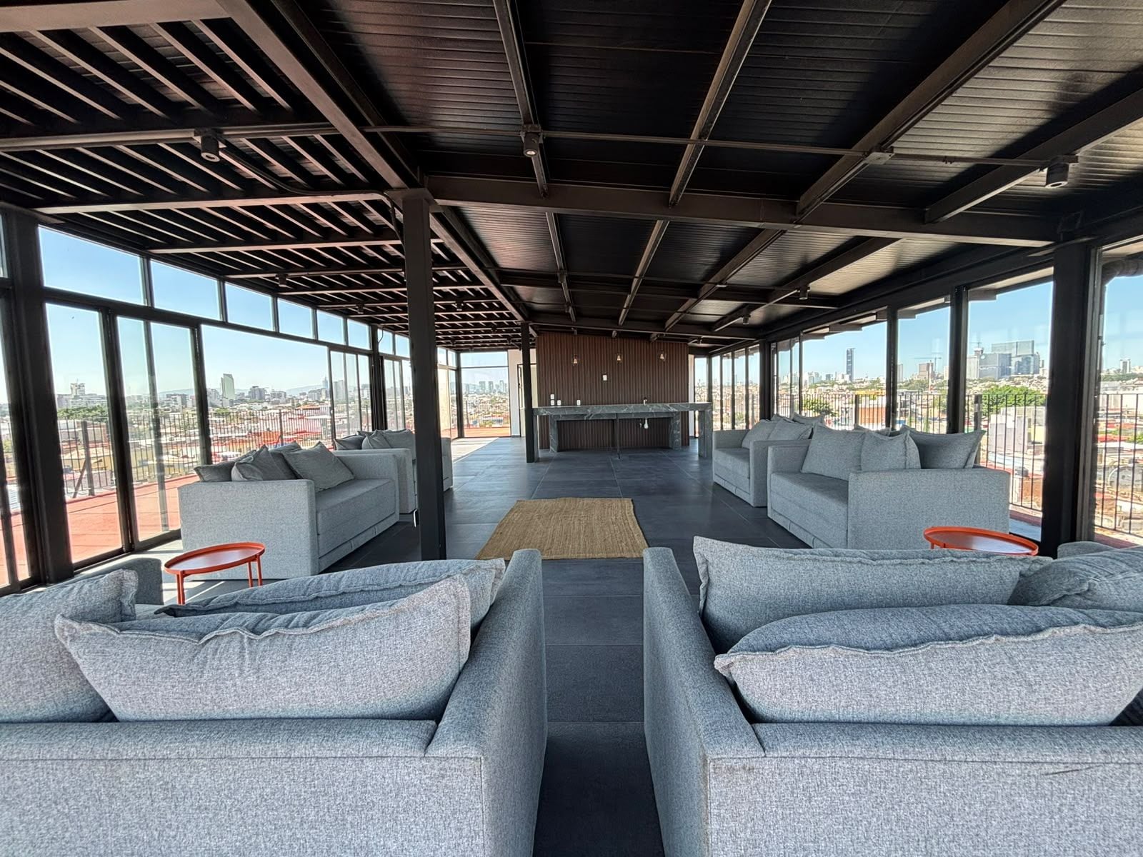 Amplia sala de estar con sofás grises y una barra, ventanas grandes con vistas a la ciudad.