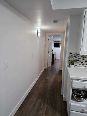 A narrow hallway with a wooden floor, white walls, and a kitchen area with white cabinets and a tiled backsplash.