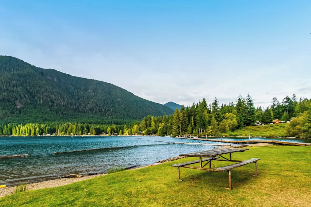A lakeside picnic area with a wooden bench and a view of a forested mountain range.