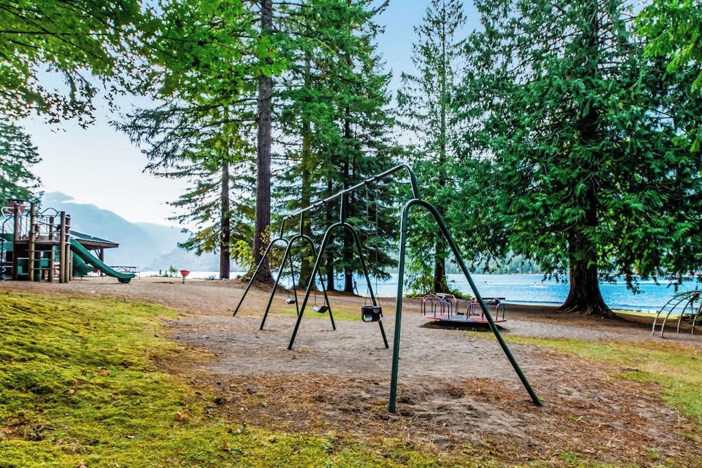 Un terrain de jeux avec des balançoires et un toboggan, entouré d’arbres et bordé par un plan d’eau.