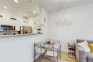 A modern kitchen with white cabinets, a microwave, and a refrigerator. There is a dining table with chairs and a small side table. A gray sofa and a decorative wall piece are also visible. - 1BR Modern Belltown Condo with Free Parking (Seattle)