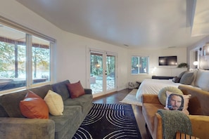 Ein modernes Wohnzimmer mit einer Couch, einem Couchtisch und einem Fernseher. Große Fenster und eine Tür führen in einen Außenbereich.