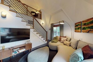 Ein modernes Wohnzimmer mit einem Flachbildfernseher, einer Holztreppe, einer runden Sitzecke und einem großen Fenster.
