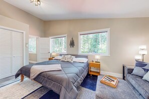 A bedroom with a bed, bedside tables, a bench, and a view of trees through the windows. - 2BR riverfront gem with kayaks & hot tub - dogs ok (Tahuya)