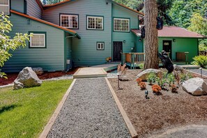 A two-story house with a front porch, a gravel driveway, and a landscaped garden with rocks and plants. - 2BR riverfront gem with kayaks & hot tub - dogs ok (Tahuya)