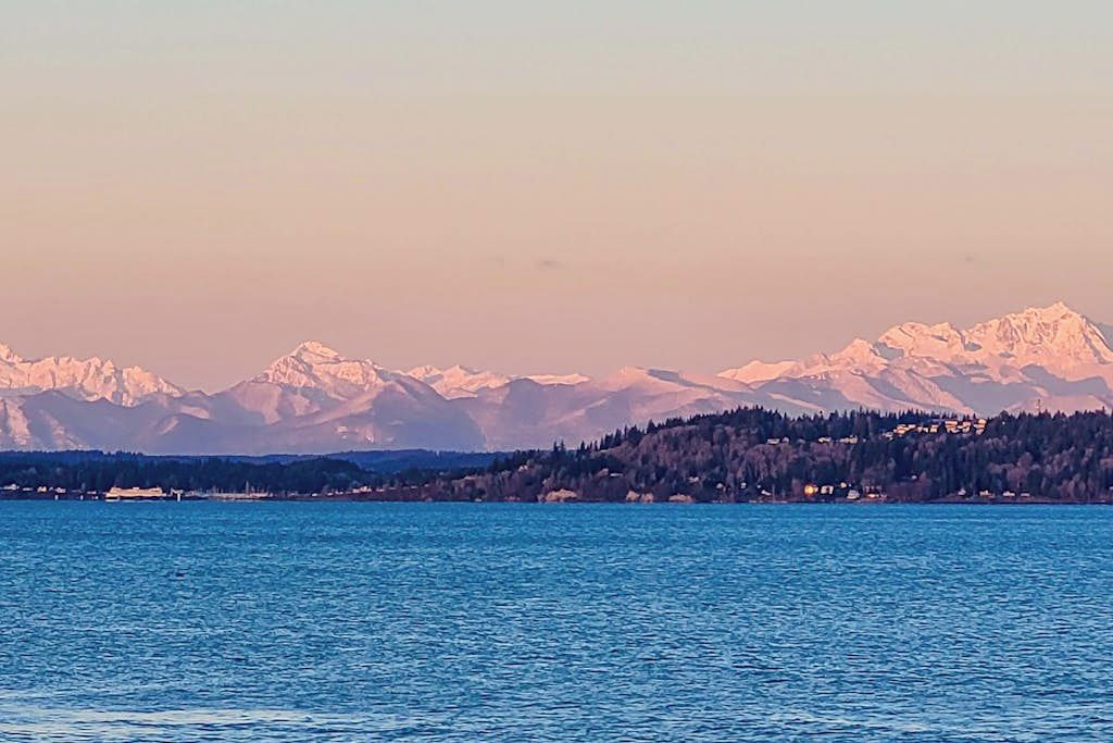 A lake with mountains in the background and a forested area with buildings along the shore.