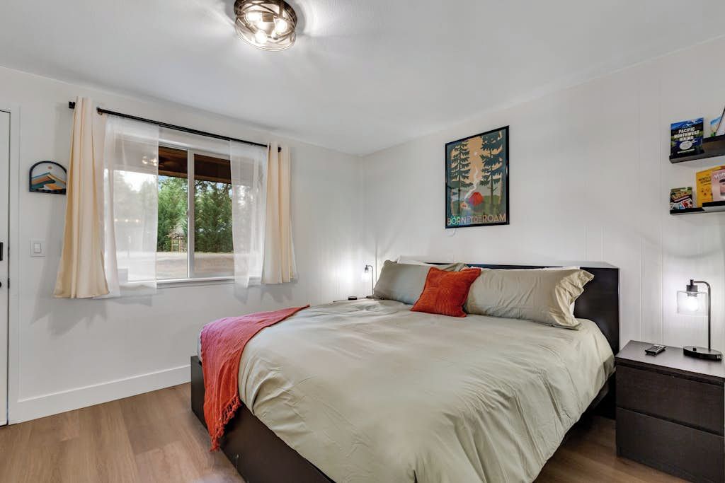 A modern bedroom with a large bed, a bedside table, a nightstand, a window with curtains, and a wall-mounted artwork.