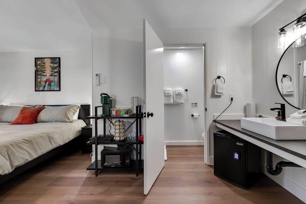 Quarto de hotel com cama, pequeno espaço de cozinha, banheiro com espelho e porta-toalhas, e porta que dá para outro quarto.
