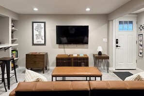 A living room with a flat-screen TV, a wooden cabinet, a bench, and a door with a window. - Two BR duplex with wifi, Washer Dryer & convenient location (Seattle)