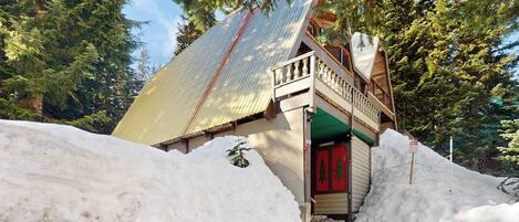 A snow-covered building with a red door, surrounded by trees and a large pile of snow.