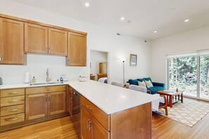 A modern kitchen with wooden cabinets and a white countertop, a living area with a blue sofa and a coffee table, and a large window offering a view of trees. - Charming 1BR Retreat - Patio - Golf Nearby - WIFI (Bainbridge Island)