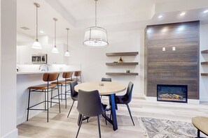 Ein moderner Essbereich mit einem runden Tisch, Stühlen, Pendelleuchten, einem eingebauten Kamin und einer Küche im Hintergrund.