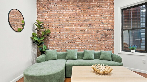 A room with a brick wall, a green sectional sofa, a round mirror, a potted plant, and a wooden coffee table.