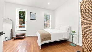 A bedroom with a bed, two windows, a wooden floor, a small table, and a wall with a geometric pattern.