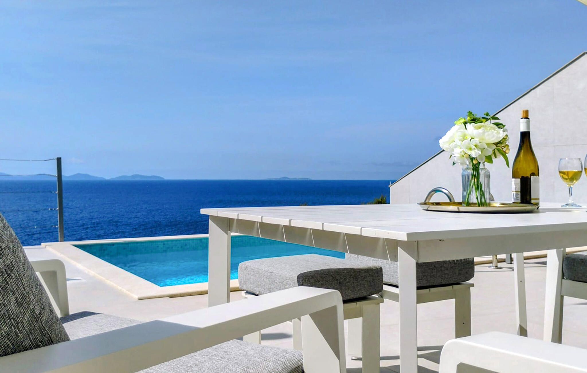 A poolside area with a view of the sea, a table with a bottle of wine and a glass, and a vase with flowers.