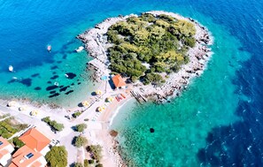 Luftaufnahme einer Küstenregion mit einer kleinen Insel, die einen Strand, einige Gebäude und klares, blaues Wasser zeigt.
