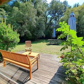 Een houten terras met een bankje en tafel, omgeven door weelderig groen en bomen.