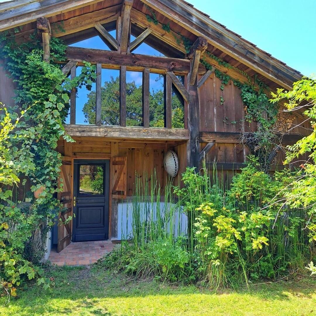 A rustic wooden house with a prominent front door, a balcony, and a garden area with various plants.