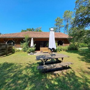 Een rustiek houten gebouw met een pannendak, een houten bank en een picknicktafel met een witte parasol.