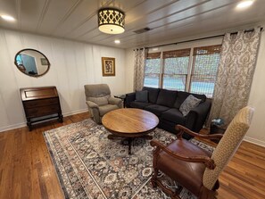 A living room with a round wooden coffee table, a dark-colored sofa, a patterned rug, a wooden chest of drawers, and a large window with patterned curtains. - 2 miles to Toomer's Corner - FREE PARKING 1 mile from Campus - AUBURN RETREAT (Auburn)