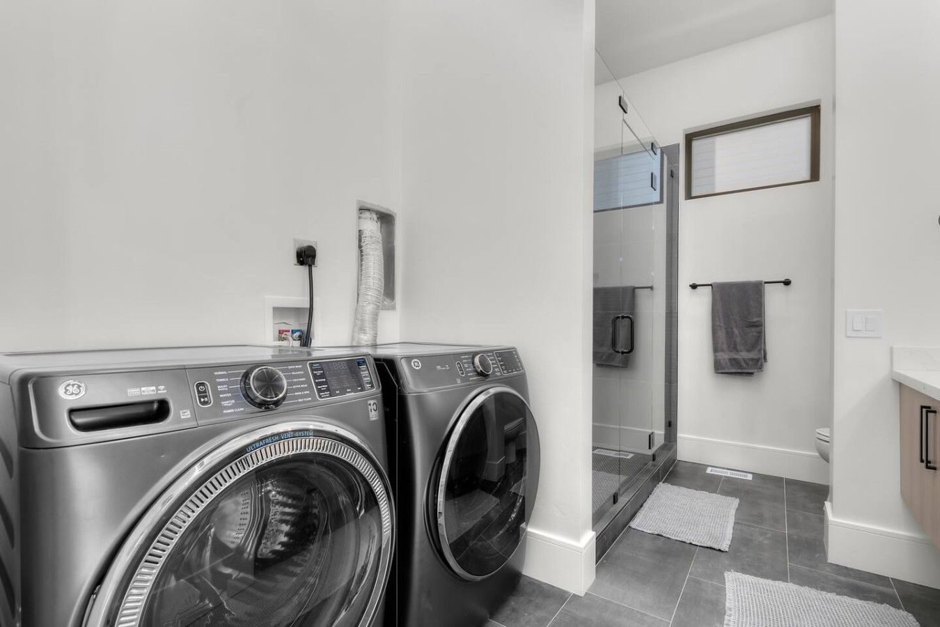 Un espace buanderie comprenant deux machines à laver à chargement frontal, une douche en verre et un sol carrelé de couleur blanche.