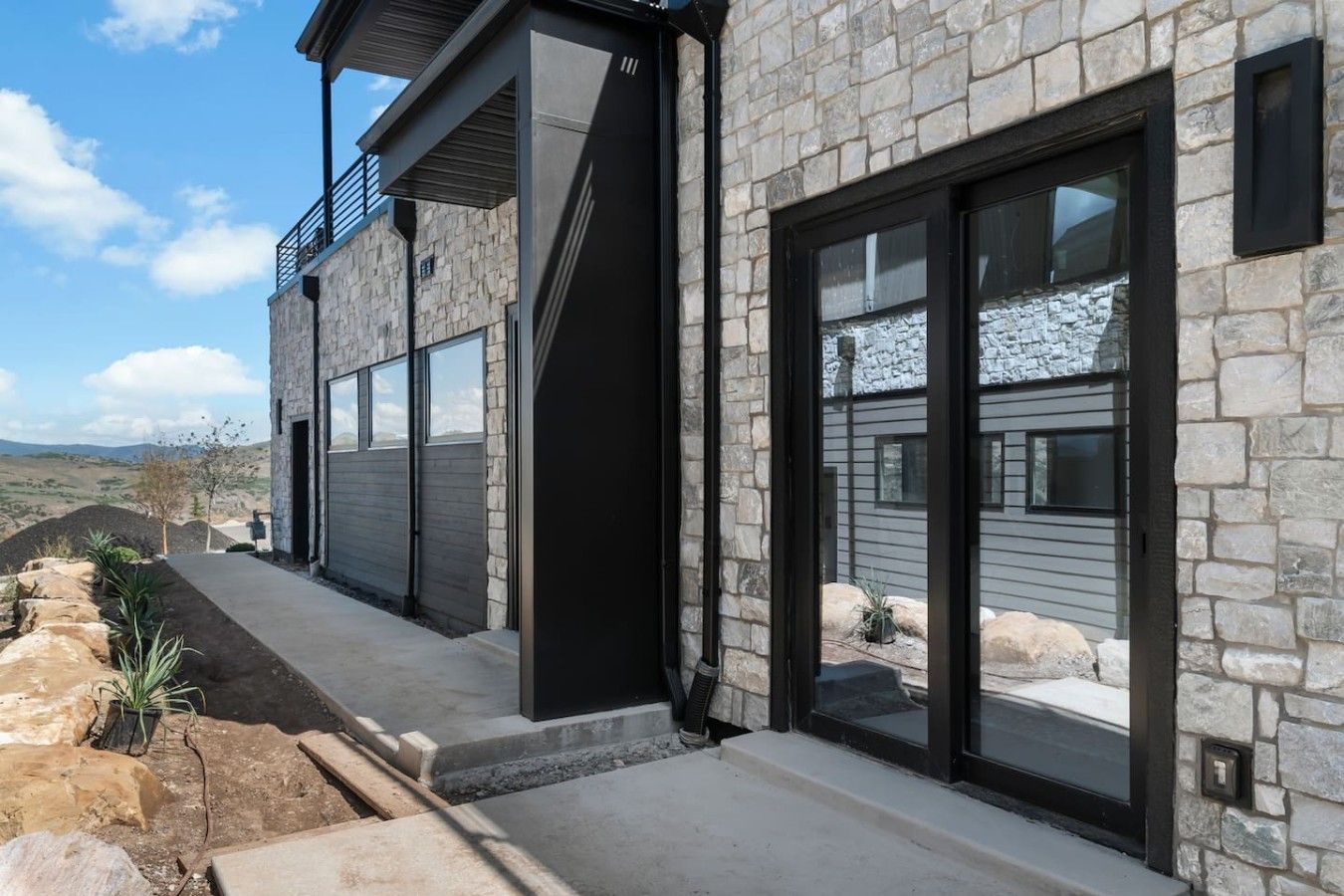 A modern two-story house with stone exterior walls, large sliding glass doors, and a balcony.