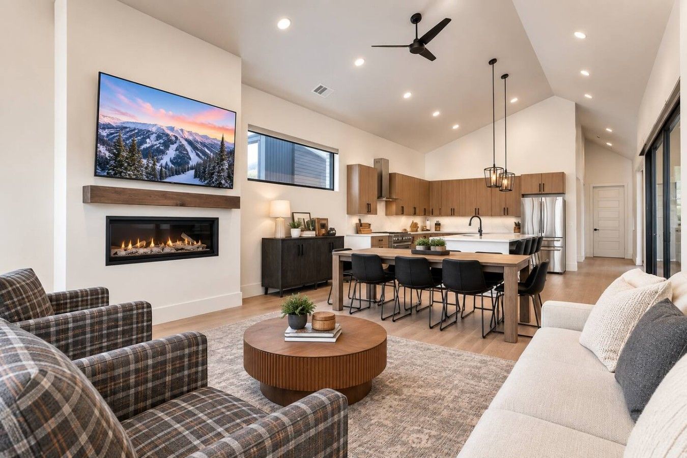 A modern living room with a fireplace, a large TV, and a dining area with a kitchen in the background.