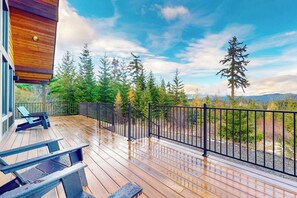 A wooden deck with a metal railing, overlooking a forested area with evergreen trees. - 5 BR modern mountain home with mountain views (Cle Elum)