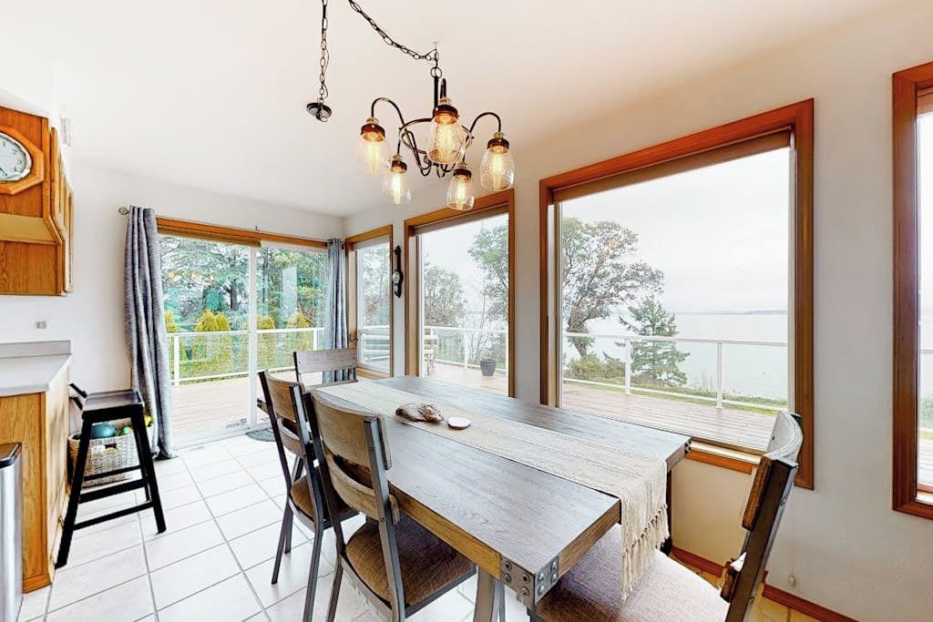 A dining area with a wooden table and chairs, large windows, and a chandelier.