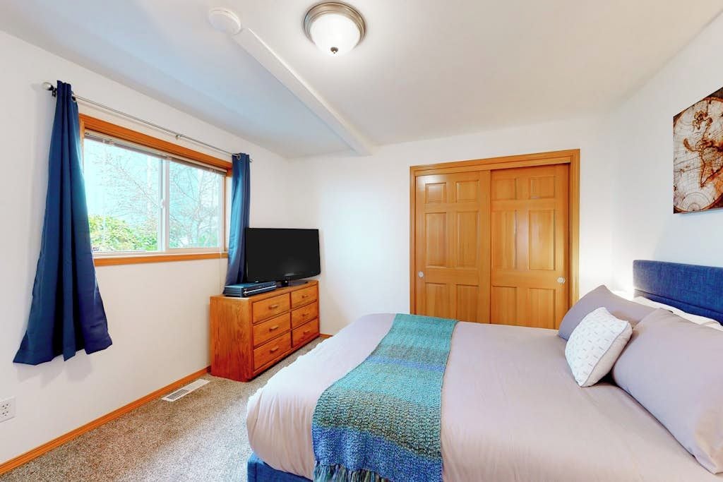 A bedroom with a bed, a television on a wooden cabinet, a window with curtains, and a door.
