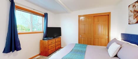 Une chambre à coucher avec un lit, un téléviseur posé sur une commode en bois, une fenêtre avec des rideaux et une porte.