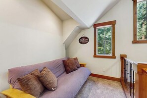 A room with a purple sofa, a 'Welcome' sign, a window with a view of trees, and a wooden staircase. - 4 BR home with private hot tub and gas fire pit (Cle Elum)